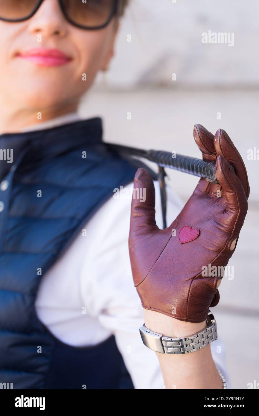 Una donna con i guanti marroni e una frusta nelle mani sta in piedi in una stalla e in primo piano Foto Stock