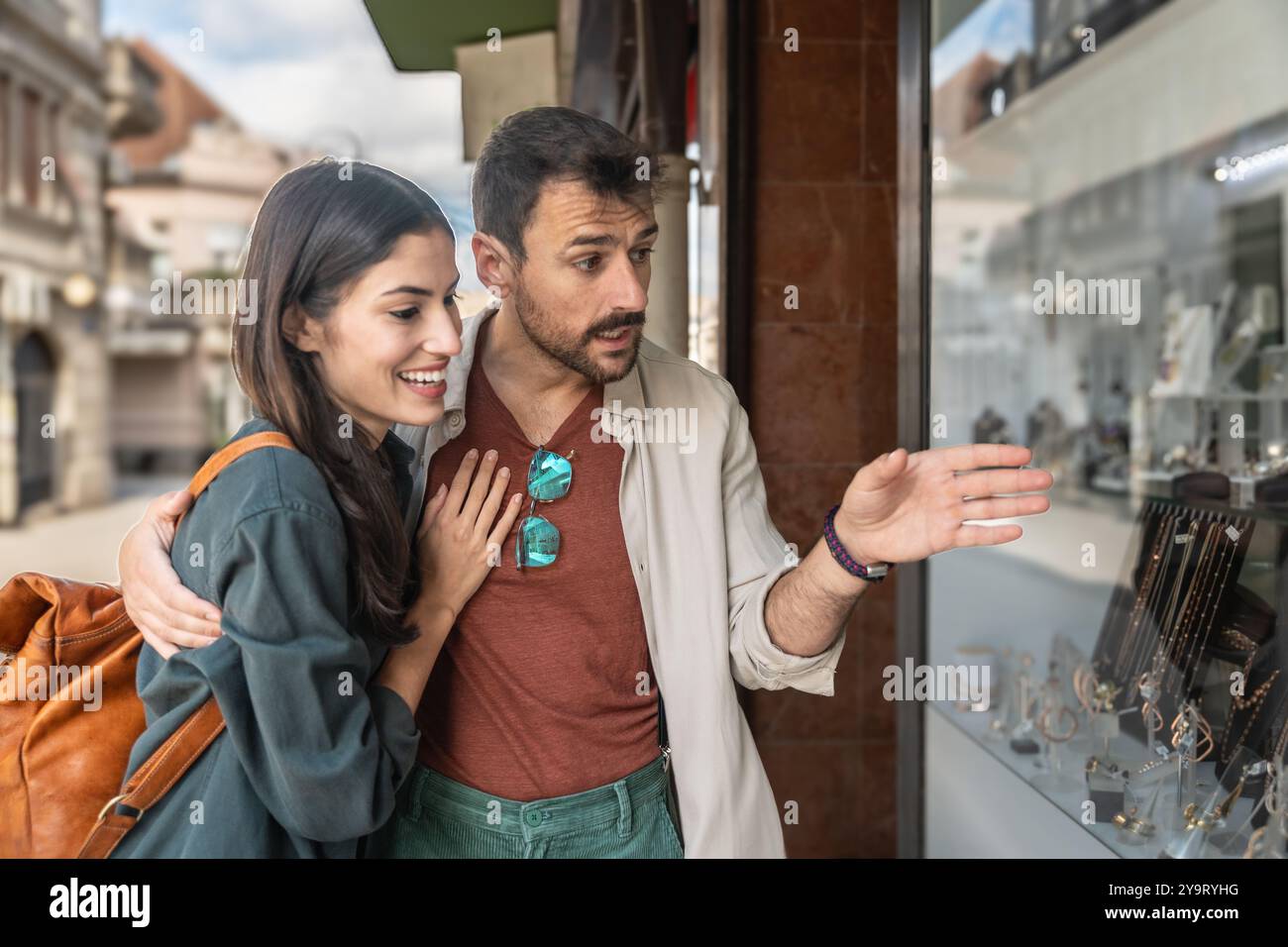 Giovane bella coppia felice innamorata in cerca di fidanzamento o di matrimonio attraverso la vetrina di vetro della gioielleria. Foto Stock