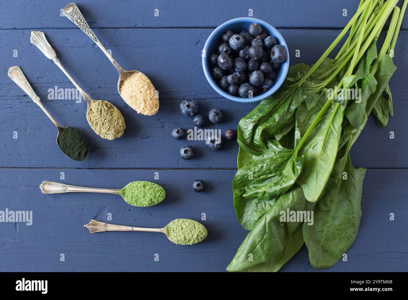 Ingredienti per frullati verdi con spirulina, erba d'orzo, proteine di canapa, spinaci e mirtilli. Immagine piatta di cucchiai antichi con polveri verdi. Foto Stock