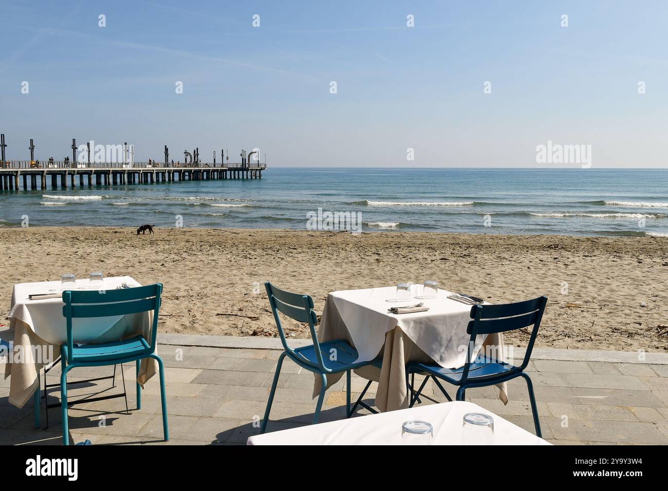 Tavoli disposti sul lungomare con la spiaggia vuota e il molo sullo sfondo in una soleggiata giornata primaverile, Alassio, Savona, Liguria, Italia Foto Stock