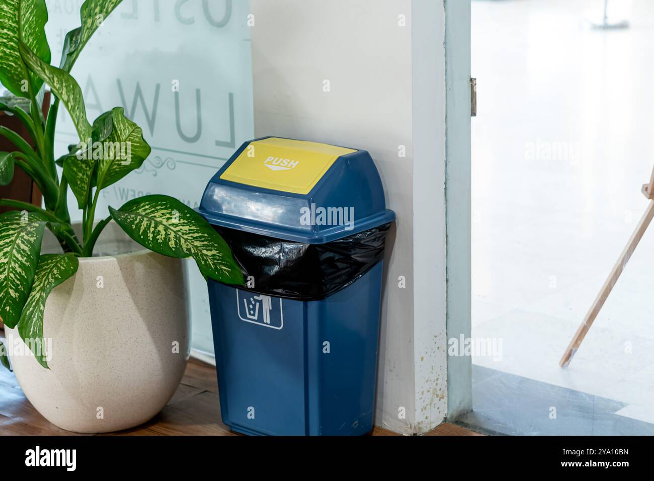 Un cestino blu con coperchio giallo, collocato accanto a una grande pianta verde in un ambiente interno moderno. Il bidone ha all'interno un sacco nero per la spazzatura ed è depositato Foto Stock