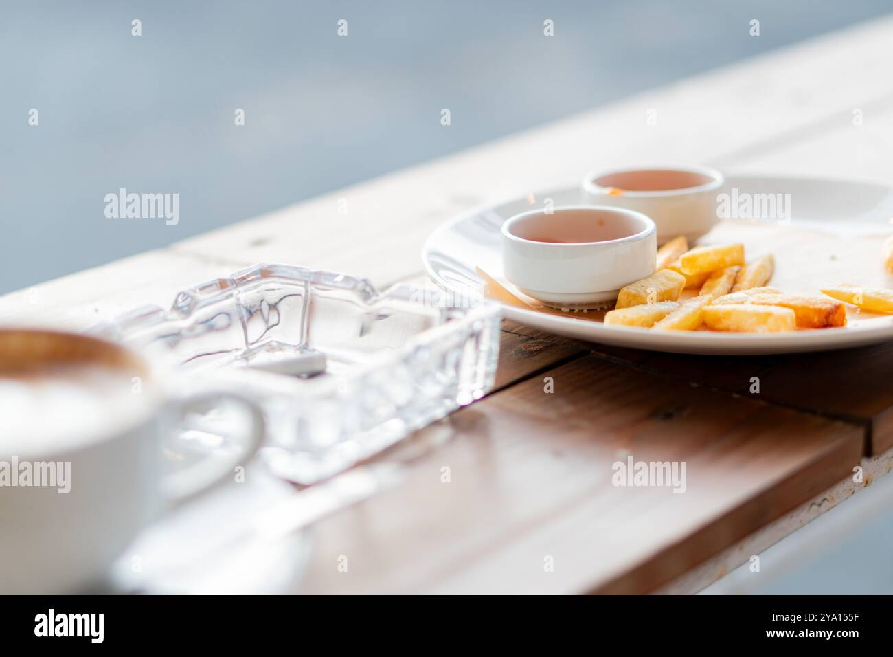 Un'accogliente caffetteria con un tavolo in legno con una tazza di caffè, un posacenere e un piatto di patatine dorate servite con salse. Lo sfondo è Foto Stock