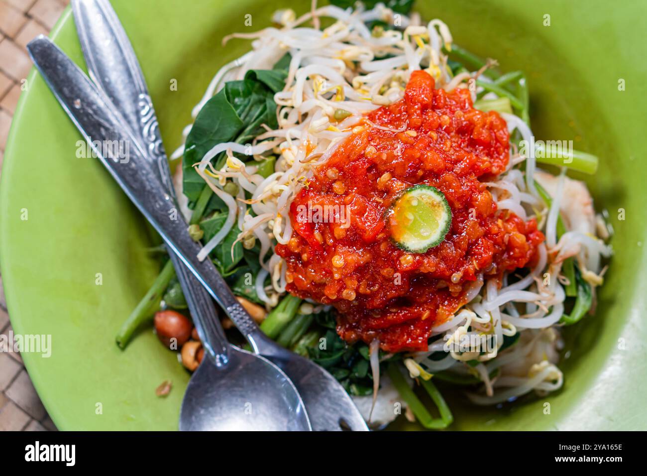 Una vivace ciotola di spaghetti condita con una salsa rossa piccante, germogli di soia freschi e verdure a foglia verde. Il piatto è guarnito con una fetta di lime e Foto Stock