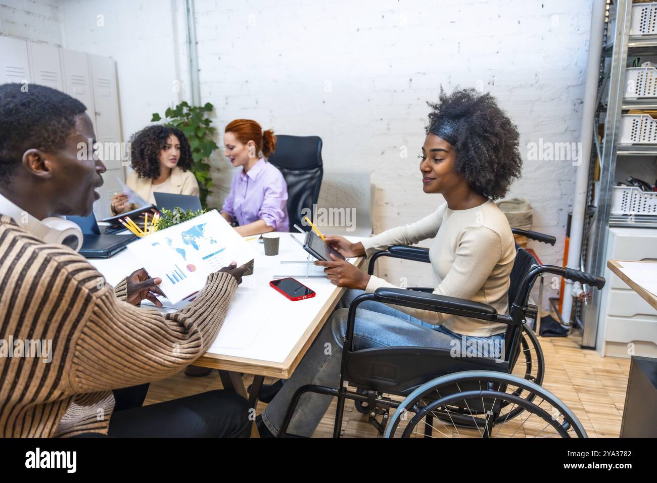 Vista laterale di una allegra donna africana con disabilità seduta su sedia a rotelle e di un collega africano maschio che lavora in un coworking Foto Stock