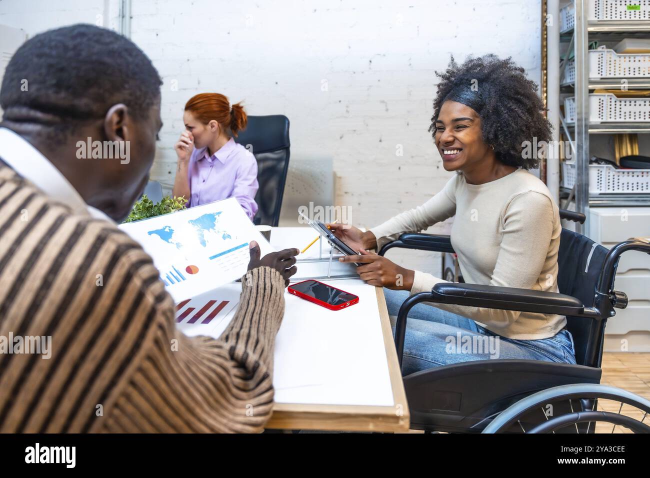 Donna africana allegra e sorridente con disabilità seduta su sedia a rotelle e collega che discute grafici in un coworking Foto Stock