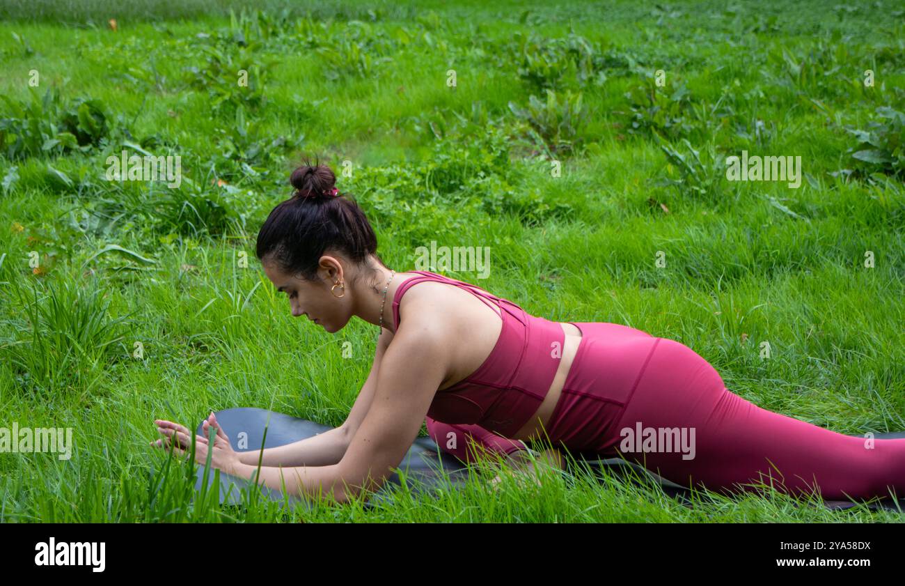 La giovane donna con i capelli bruna tirata indietro in un panino esegue una posa yoga per lavorare sull'apertura dell'anca vestita con un bel vestito rosso da yoga con top e leggings Foto Stock