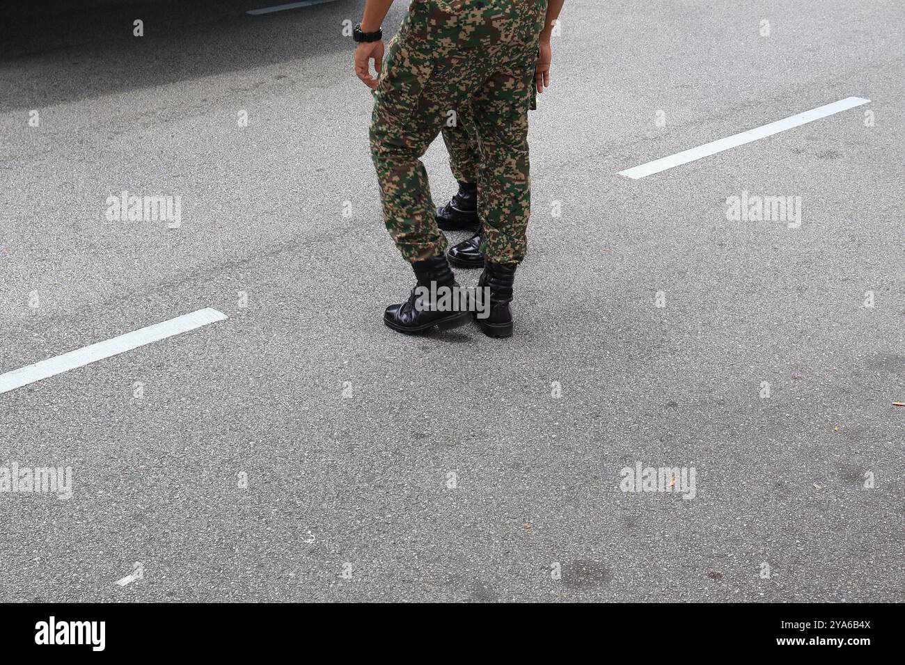 Primo piano dell'uniforme militare mimetica e degli stivali neri su una strada asfaltata, raffigurante la prontezza e la forza. Foto Stock