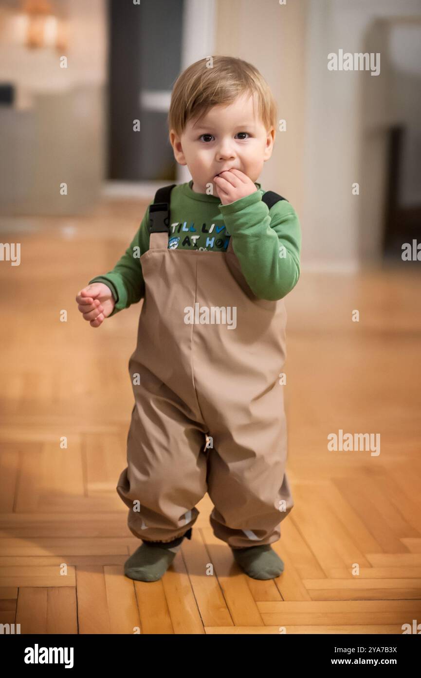 Un bambino gioioso, vestito con pantaloni da sci, si solleva con sicurezza mentre masticava giocosamente le dita in un ambiente interno accogliente. Foto Stock