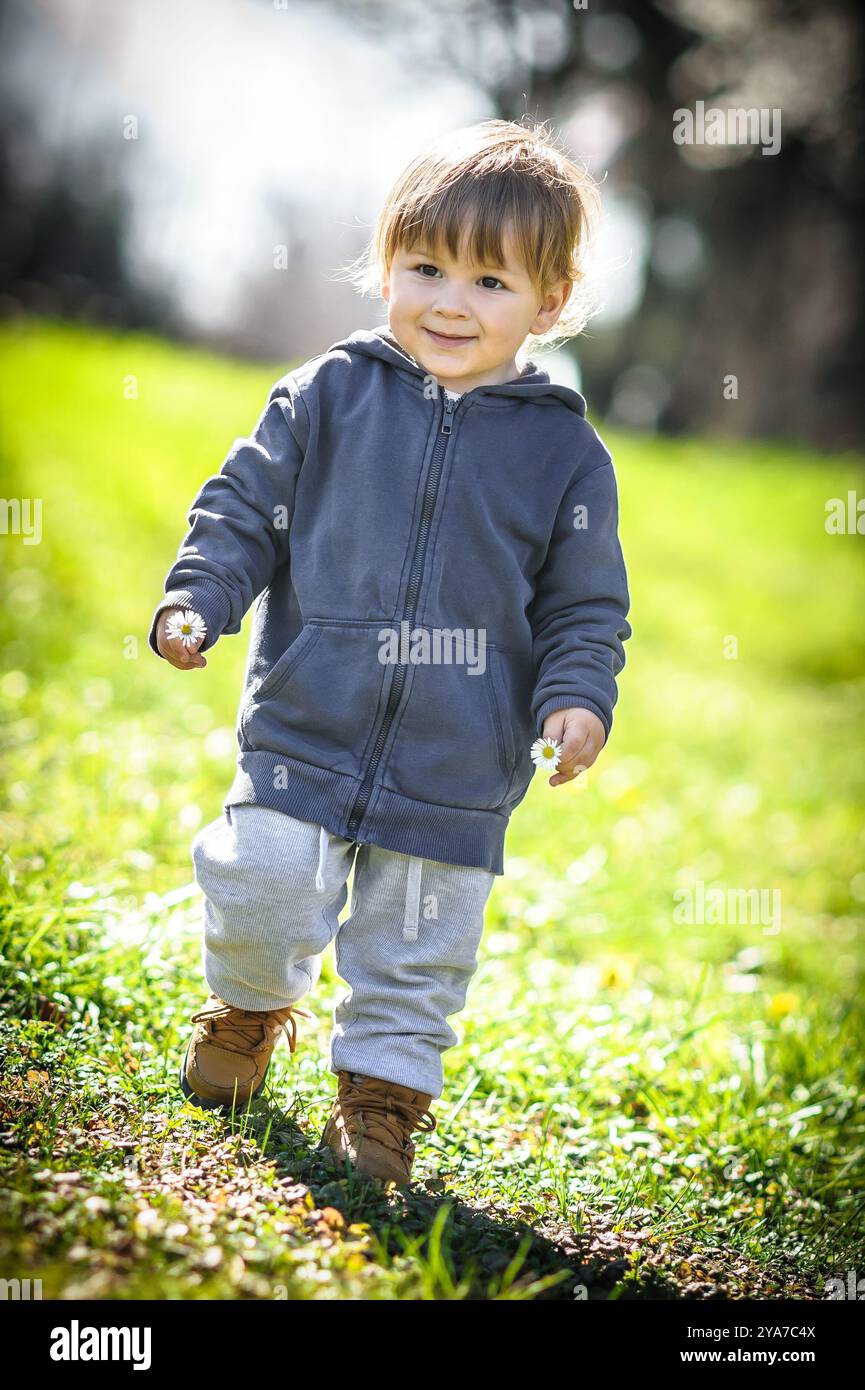 Il bambino che indossa una felpa con cappuccio e pantaloni felicemente cammina attraverso un campo verde vibrante, godendosi il sole luminoso e il clima caldo. Foto Stock