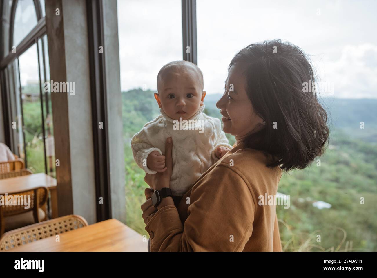 Una donna tiene delicatamente un bambino dolce tra le braccia vicino a una finestra Foto Stock