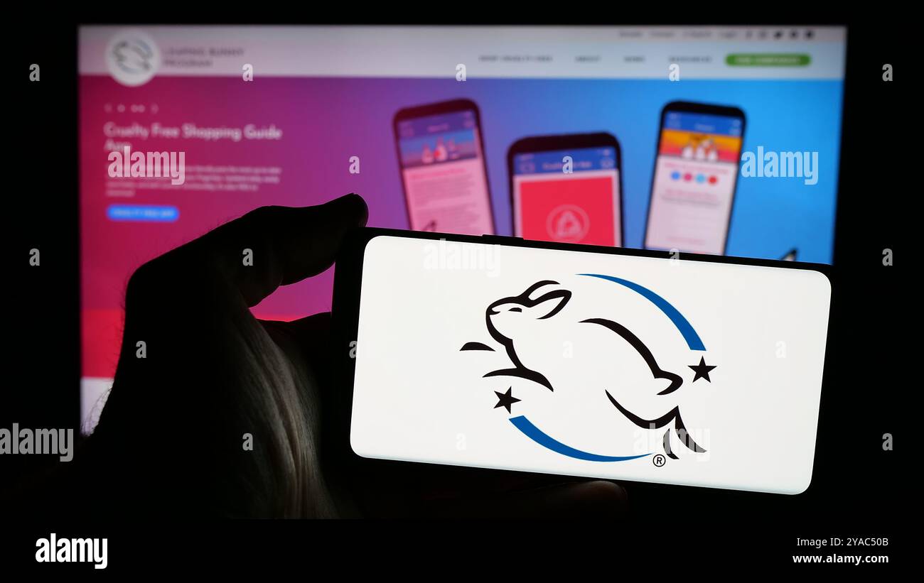 Persona che possiede un telefono cellulare con il logo dell'organizzazione di certificazione Leaping Bunny Program sullo schermo davanti alla pagina Web. Mettere a fuoco il display del telefono. Foto Stock
