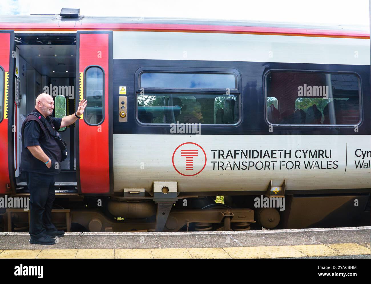 Un macchinista si alza mentre aspetta accanto a un treno in attesa in una stazione. Una signora anziana a bordo guarda fuori dalla finestra. Trasporto per il Galles Foto Stock