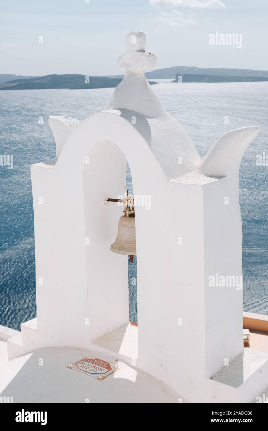 SANTORINI, GRECIA - 30 settembre 2024: Arco bianco con campana della chiesa con acqua blu sullo sfondo. Foto Stock