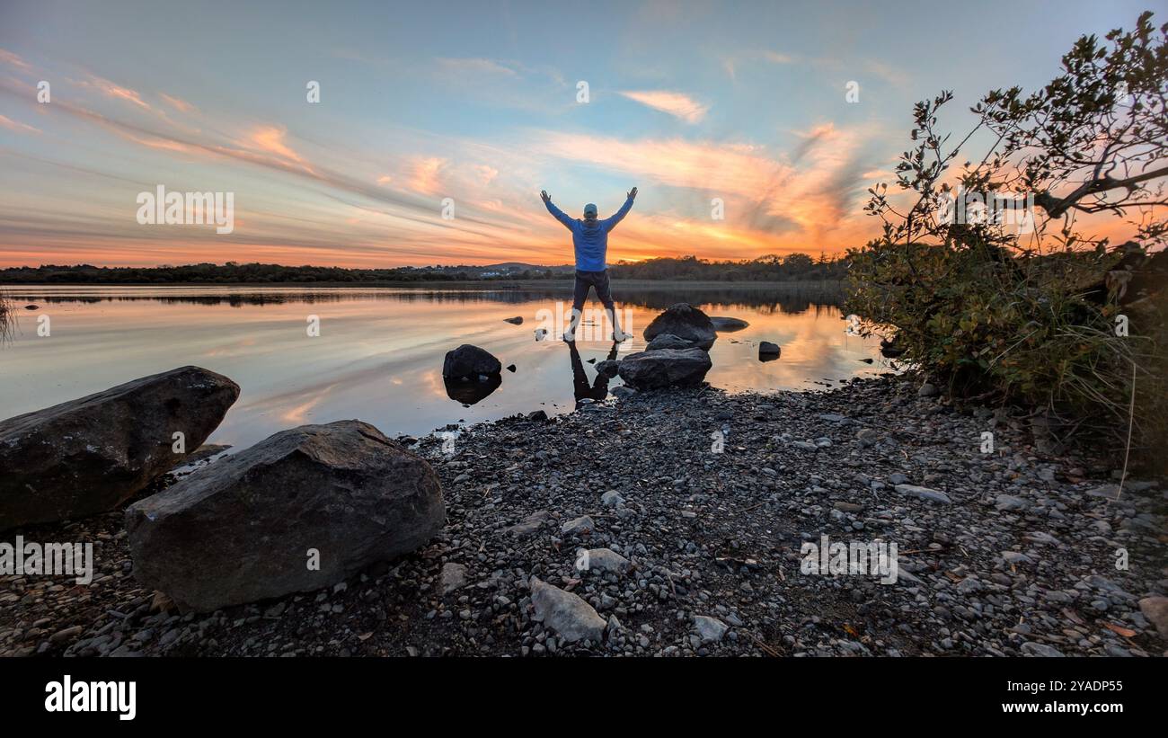 Splendido tramonto arancione sul lago, l'uomo si riflette nell'acqua a Ballyquirke lough, Galway, Irlanda, sullo sfondo naturale Foto Stock