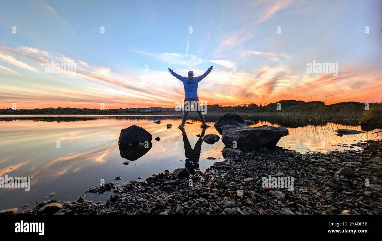 Splendido tramonto arancione sul lago, l'uomo si riflette nell'acqua a Ballyquirke lough, Galway, Irlanda, sullo sfondo naturale Foto Stock