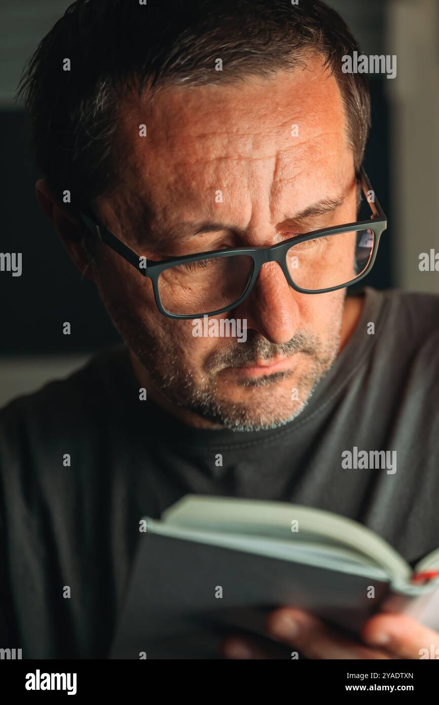 Concetto Bookworm. Lettura di un libro a casa, da solo a tarda notte, ritratto basso di un maschio adulto maturo, messa a fuoco selettiva. Foto Stock