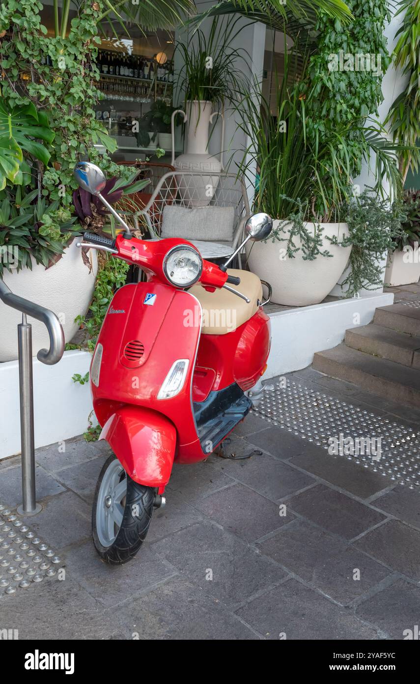 BRISBANE, QUEENSLAND, AUSTRALIA. 11 settembre 2024; Vespa rossa parcheggiata fuori da un bar decorato con foglie verdi. orientamento verticale. Foto Stock