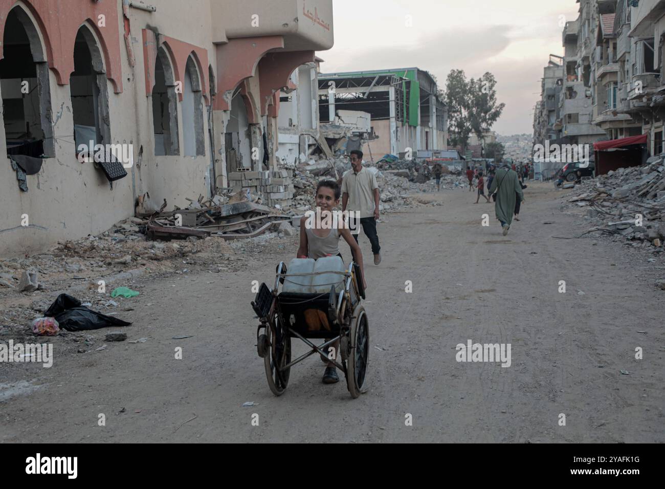 Gaza. 13 ottobre 2024. I palestinesi camminano su una strada con edifici distrutti dagli scioperi israeliani nella città meridionale della Striscia di Gaza di Khan Younis, il 13 ottobre 2024. Crediti: Rizek Abdeljawad/Xinhua/Alamy Live News Foto Stock