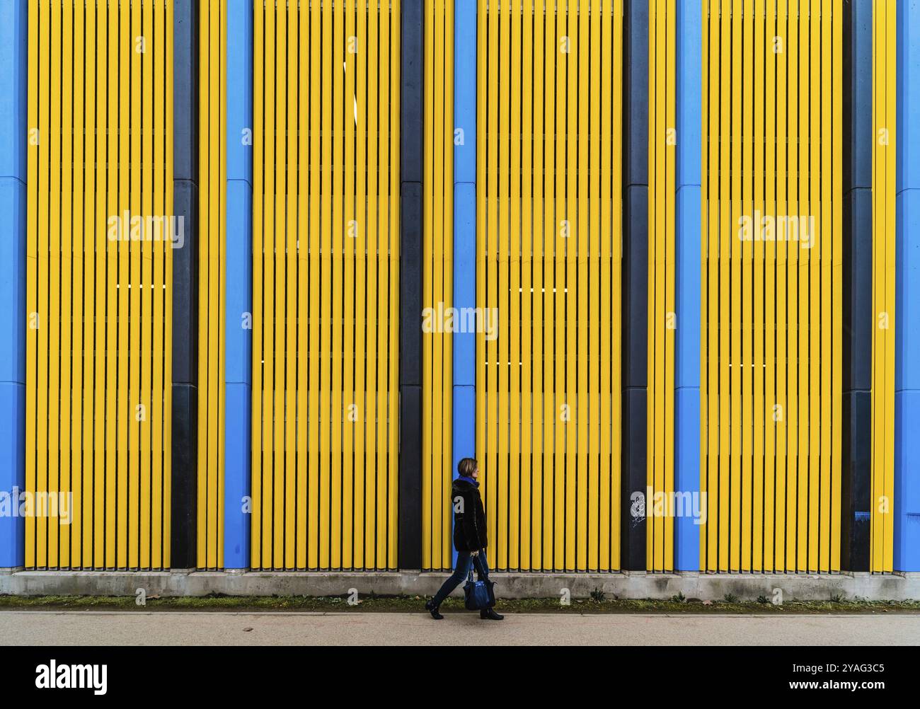 Donna pedonale che cammina di fronte a un colorato sfondo industriale con linee verticali a motivi dritti, Bruxelles, Belgio, Europa Foto Stock