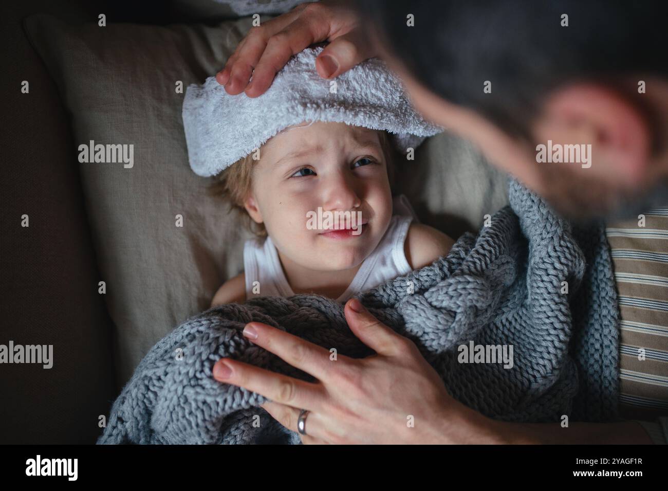 Padre che si prende cura del figlio malato con febbre e freddo, tenendo un panno freddo alla fronte. Ragazzo disteso sul divano, si sente stanco. Vista dall'alto. Foto Stock