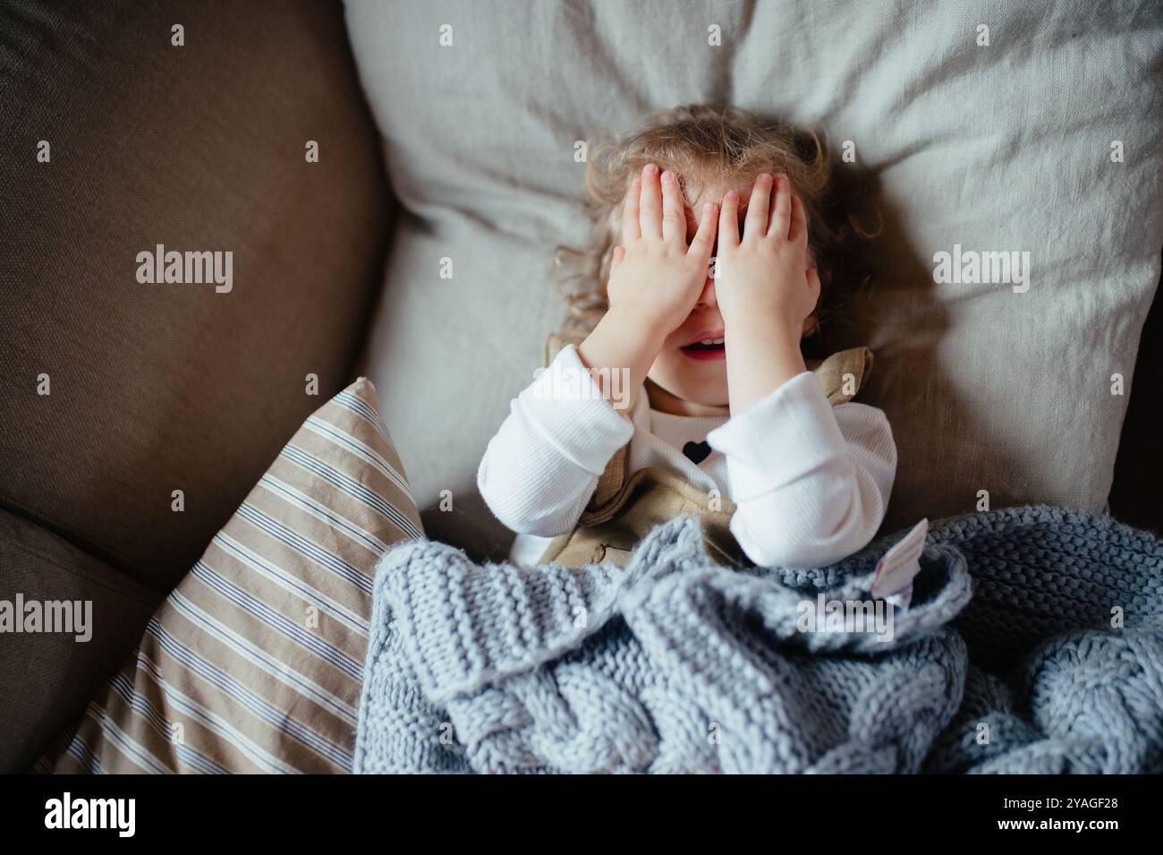 Ragazza malata con febbre e freddo sdraiata sul divano, si sente stanca, copre il viso con le mani. Foto Stock