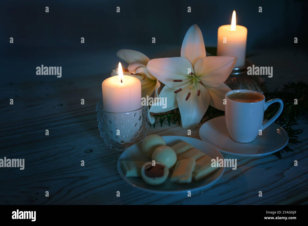 Due candele, fiori di giglio bianchi, tazza da caffè e praline, luce festosa su sfondo blu scuro, natura morta per l'Avvento, Natale e inverno Foto Stock