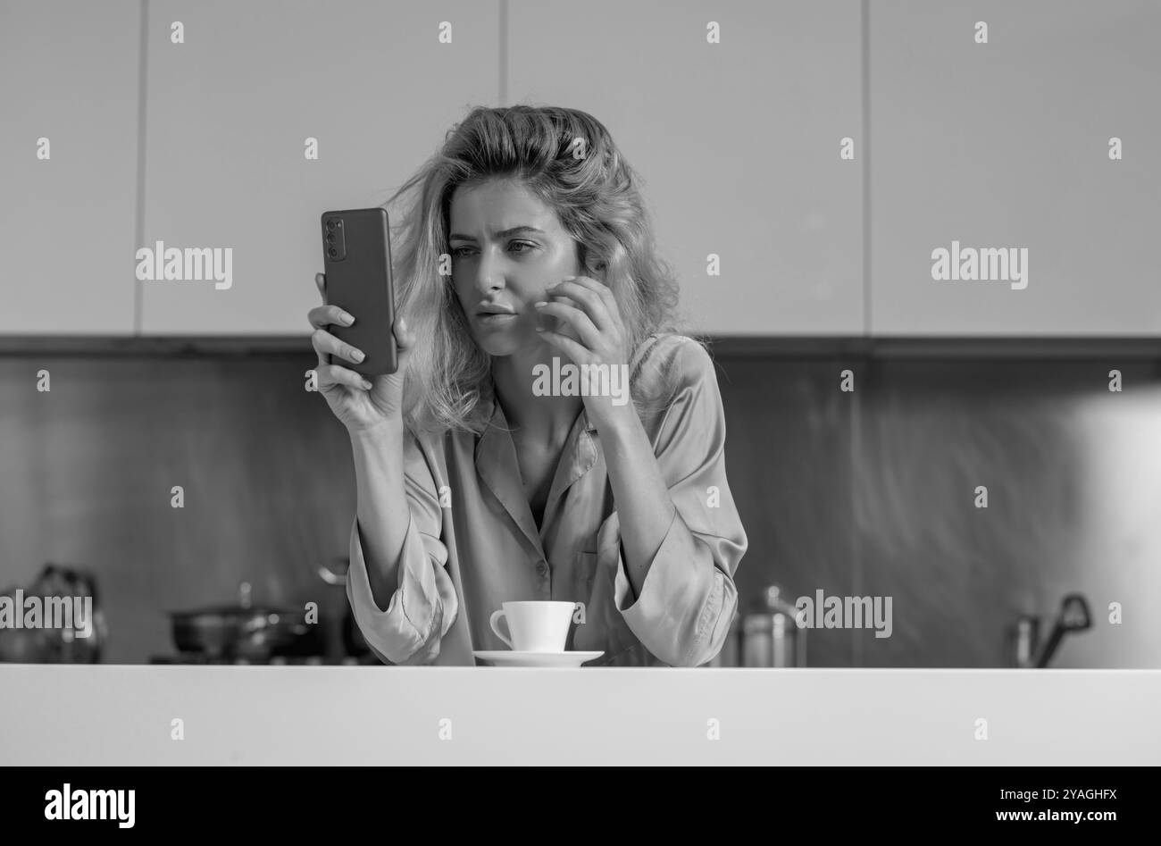 Donna stanca infelice che chiacchiera sul telefono cellulare in cucina a casa. L'umore della casalinga al mattino. Foto Stock