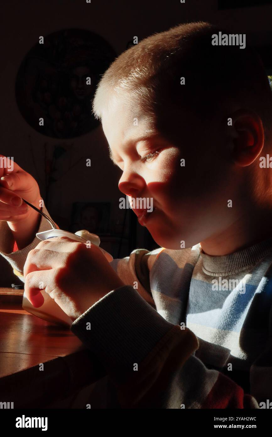 Un bambino con vitiligine mangia lo yogurt con un cucchiaio, luce dura Foto Stock