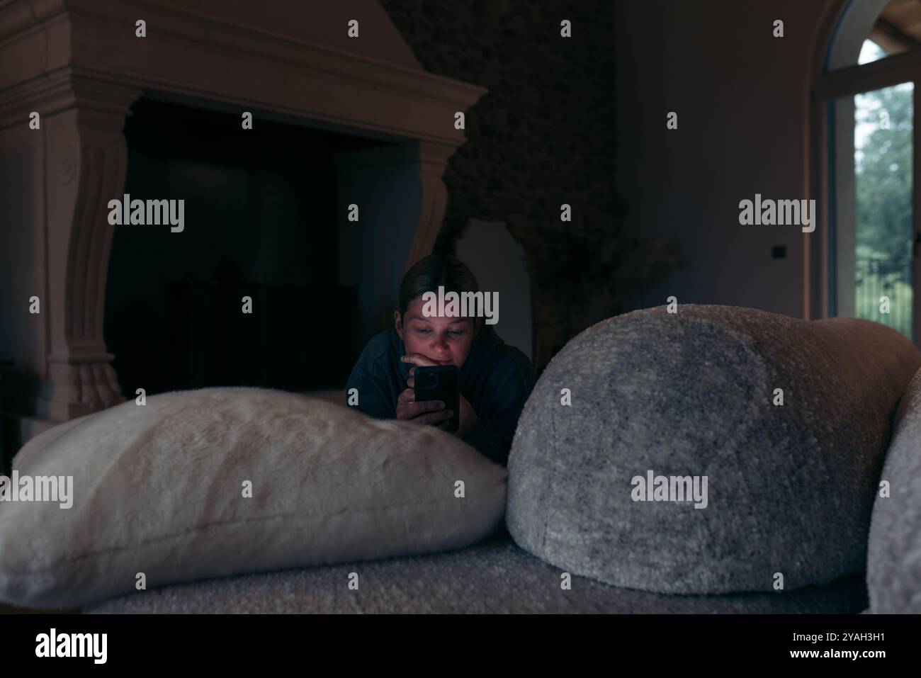 Persona sdraiata su morbidi cuscini in una stanza poco illuminata, utilizzando uno smartphone Foto Stock