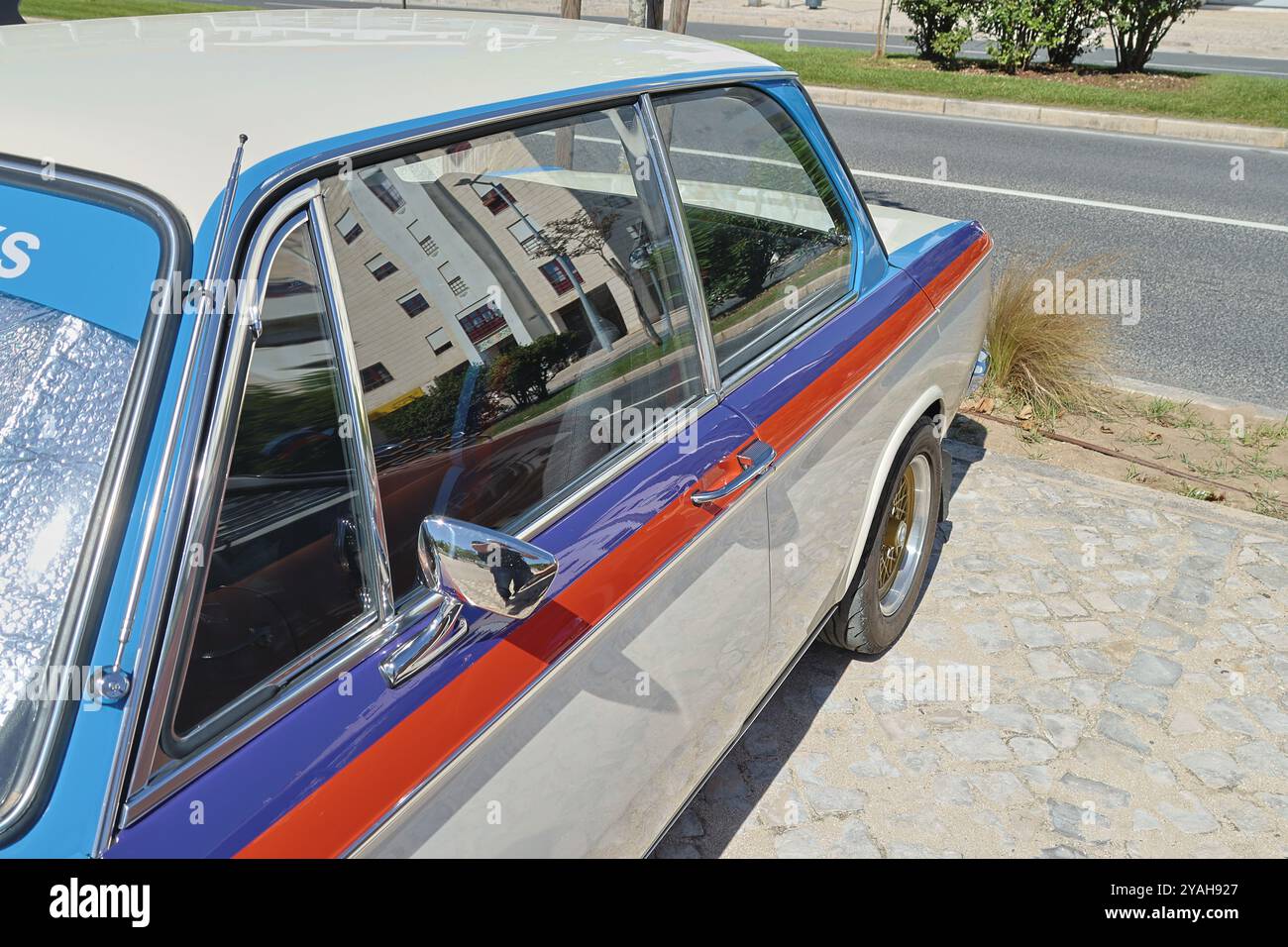La classica bmw 2002 sfoggia una livrea bianca, blu e rossa, parcheggiata su una strada acciottolata in una giornata di sole Foto Stock