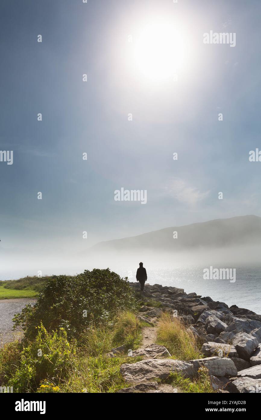 Villaggio di Ullapool, Scozia. Vista di una signora che cammina sulle rive del Loch Broom, con una nebbia di mare sullo sfondo. Foto Stock