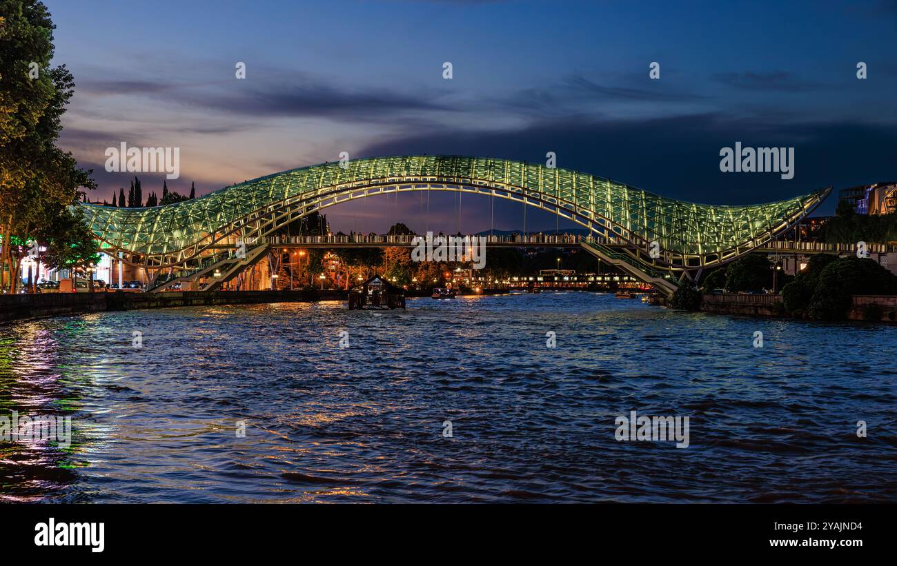 vista lungo il fiume kura a tbilisi dell'iconico ponte della pace illuminato al tramonto Foto Stock