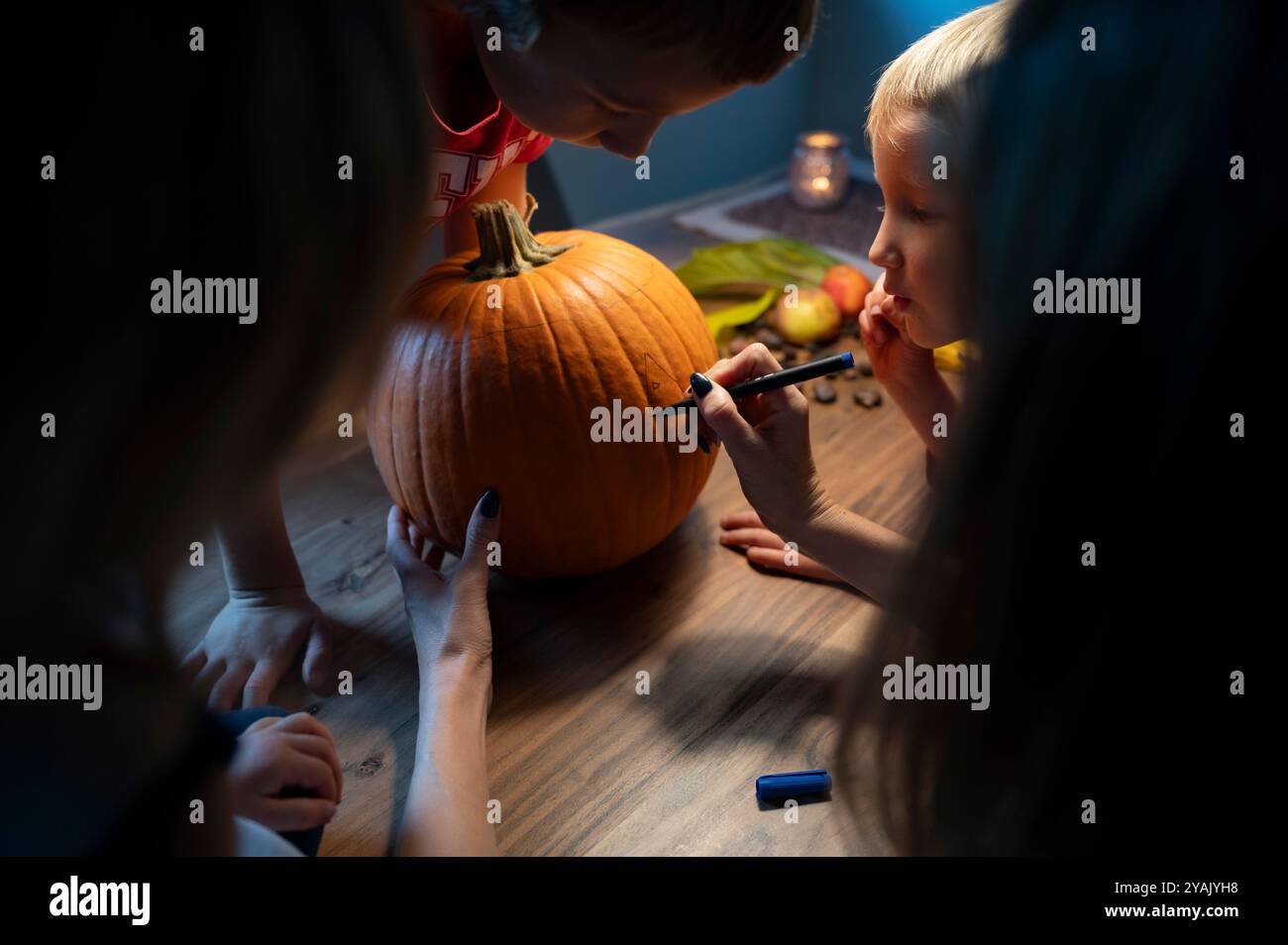 I bambini osservano attentamente mentre un adulto disegna un disegno su una zucca, preparandosi per una divertente sessione di intaglio della zucca. L'ambiente accogliente e a luce di candela lo rende ancora più piacevole Foto Stock