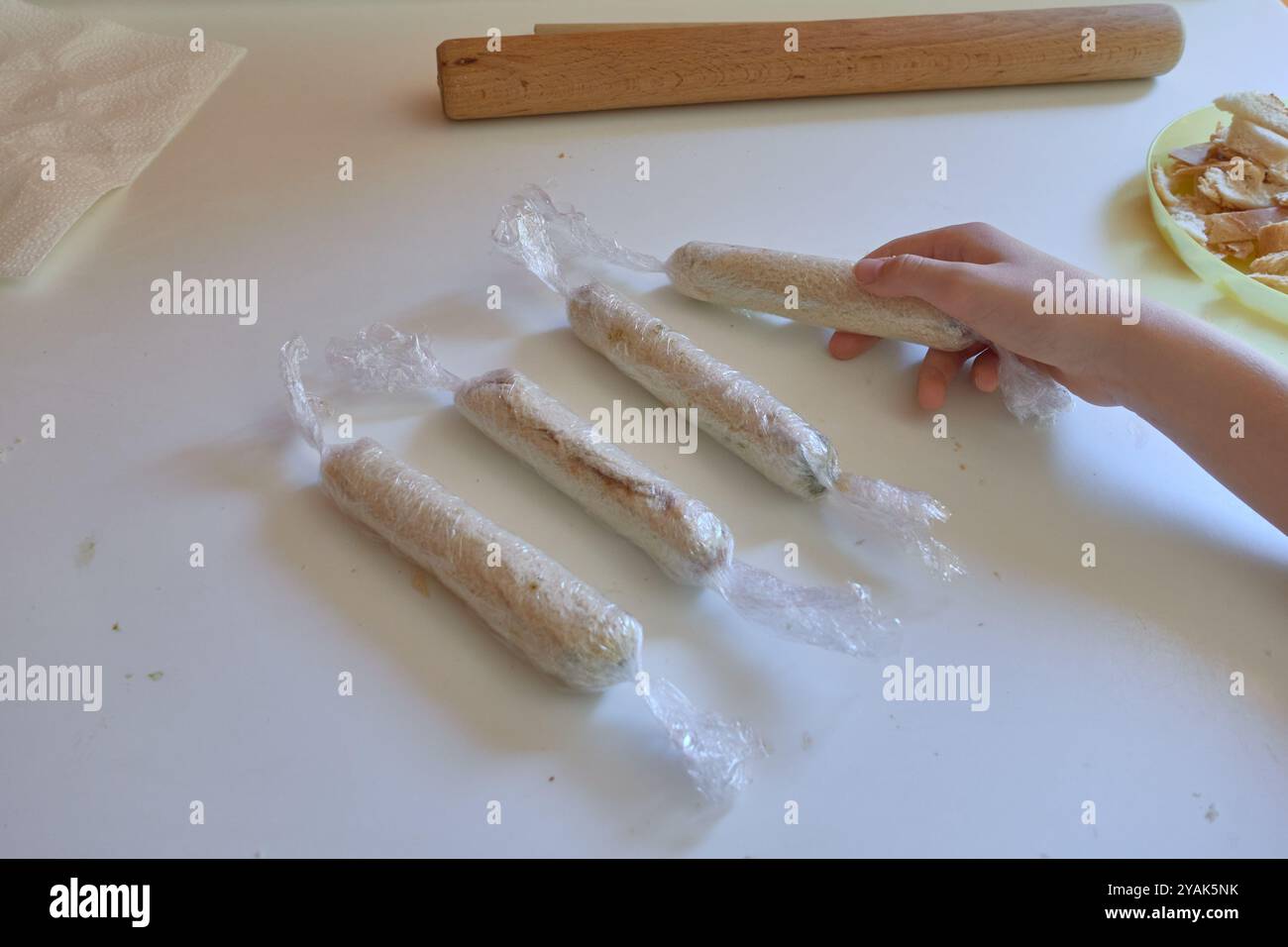 Mano femminile che prende un rotolo da una fetta di pane riempito di cioccolato avvolto in pellicola trasparente per preparare involtini alla cannella fatti in casa Foto Stock