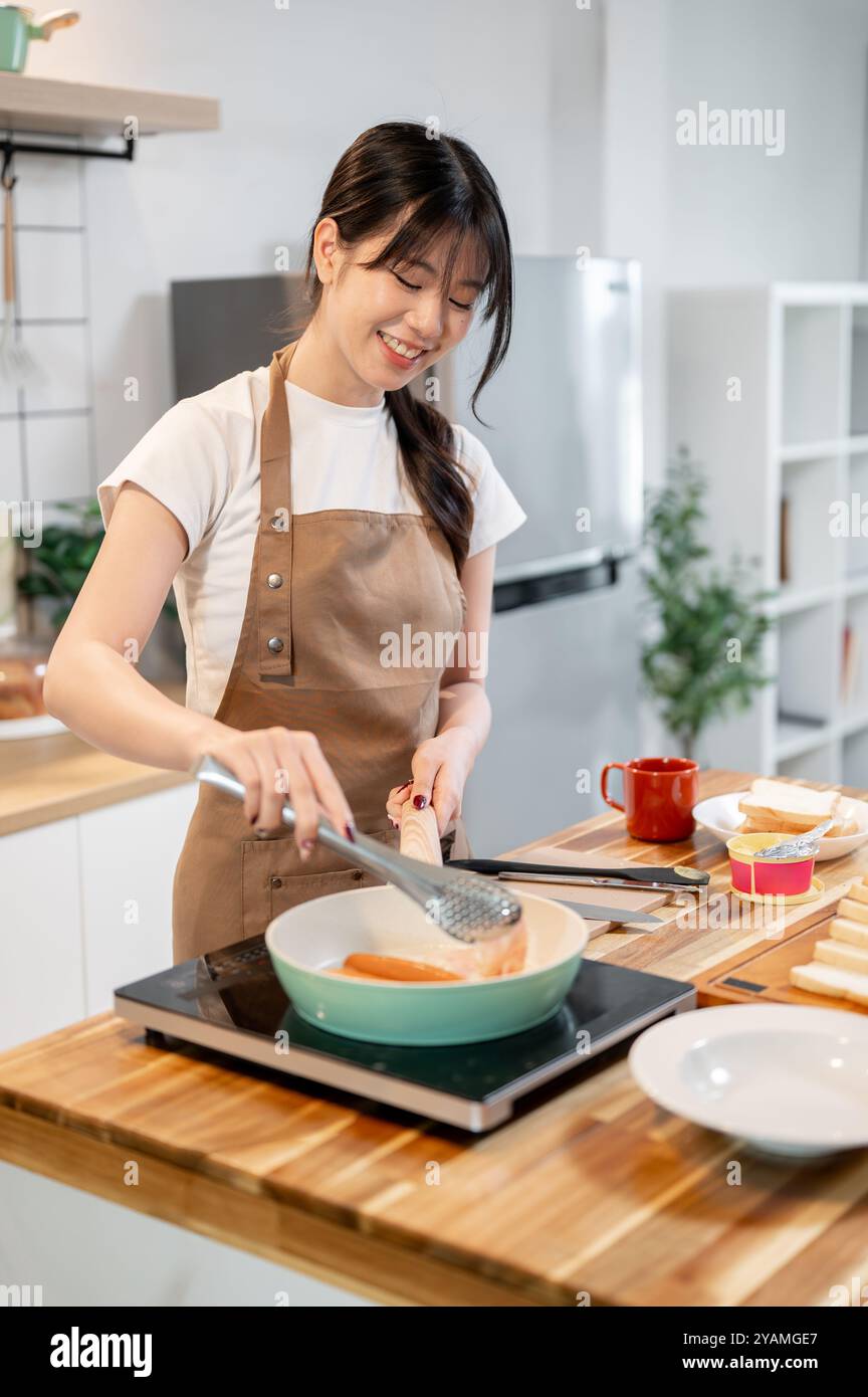 Una bella e felice donna asiatica in un grembiule si sta gustando la colazione cucinata in cucina, fritgendo salsicce in una padella. cucina casalinga, vita domestica, gente Foto Stock