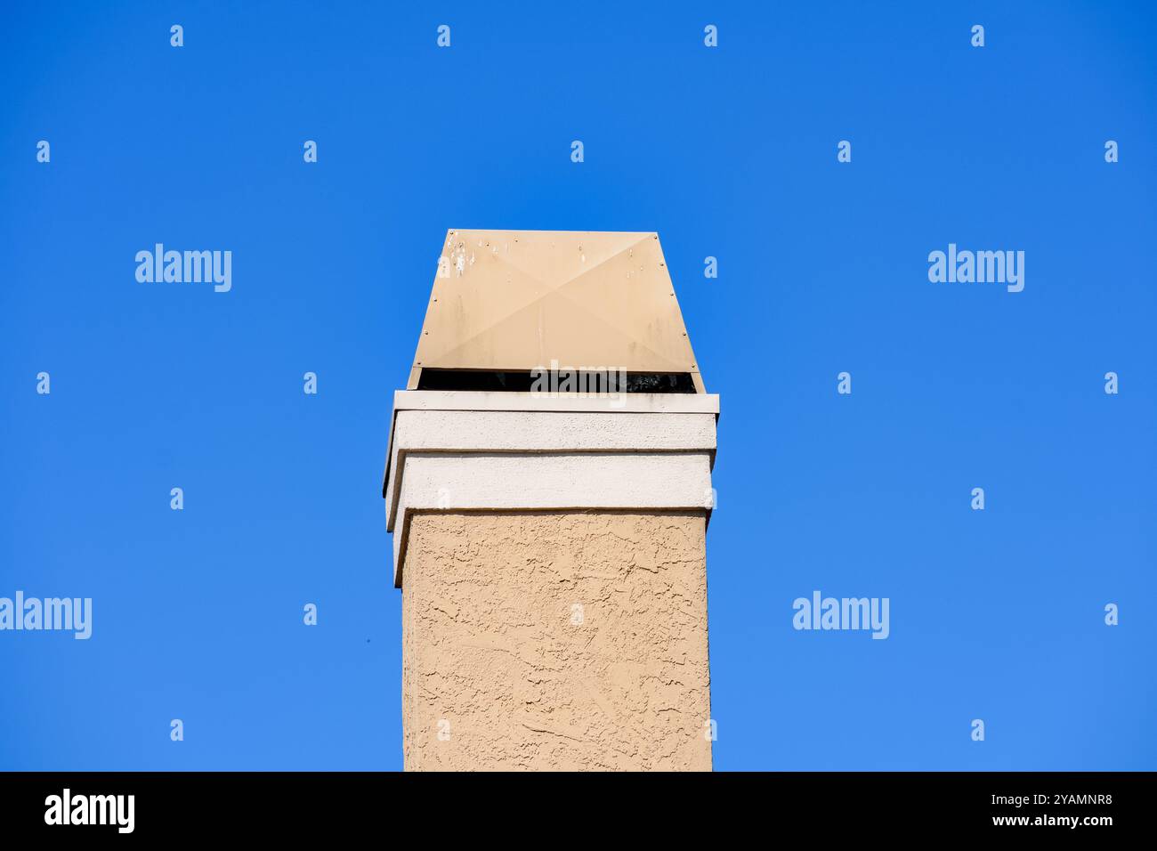 Un camino beige in stucco con coperchio di ventilazione in metallo si erge contro un cielo blu brillante. Il coperchio della bocchetta di ventilazione mostra segni di usura. Foto Stock