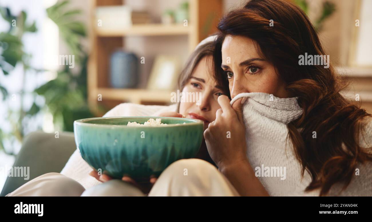 Spaventosa, madre matura con la figlia sul divano per legarsi, programmi televisivi o popcorn nel soggiorno. Weekend, streaming e mamma sul divano con Foto Stock