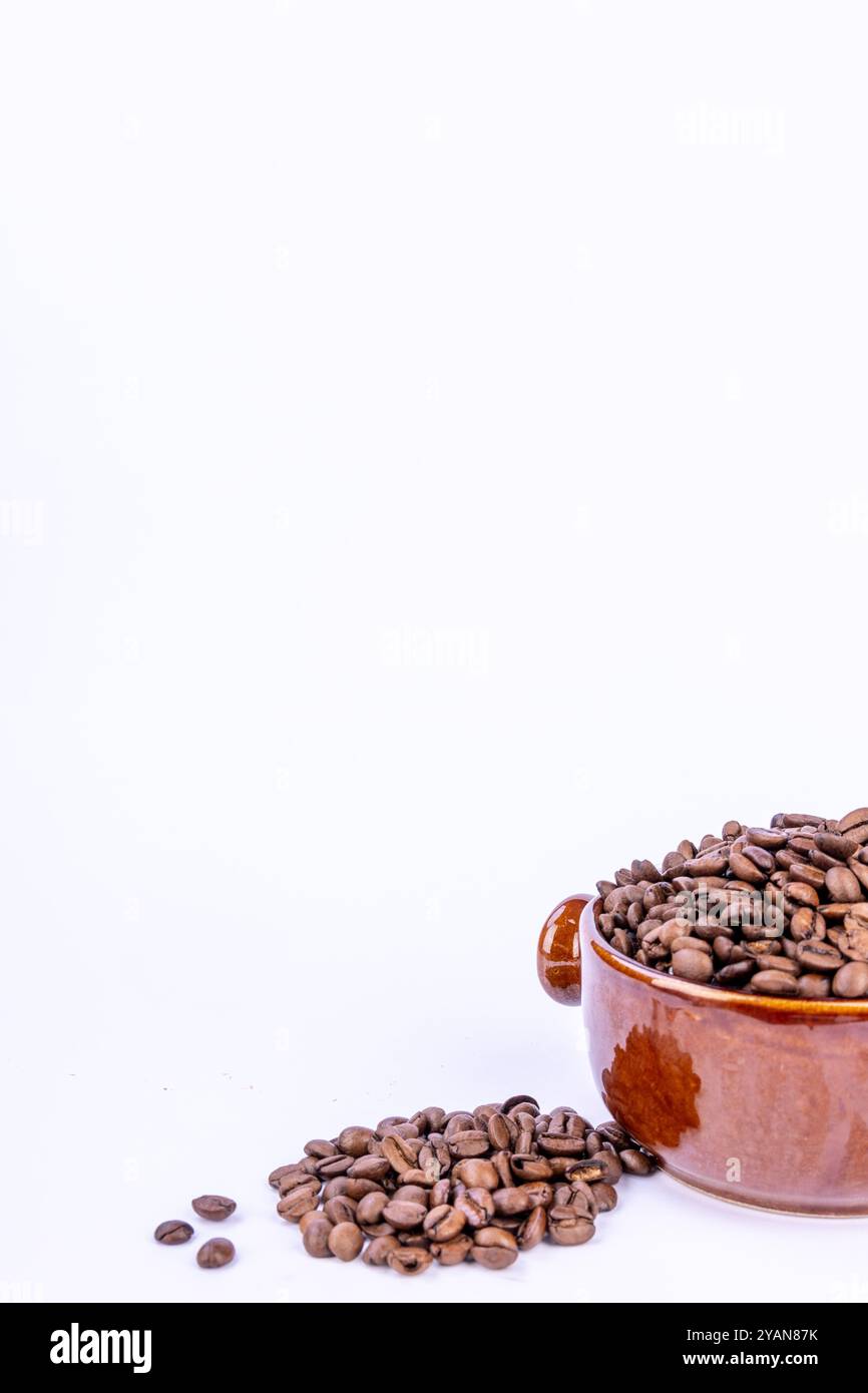 Una ciotola marrone calda piena di chicchi di caffè naturali, con pochi chicchi sparsi intorno, il tutto su uno sfondo bianco brillante Foto Stock