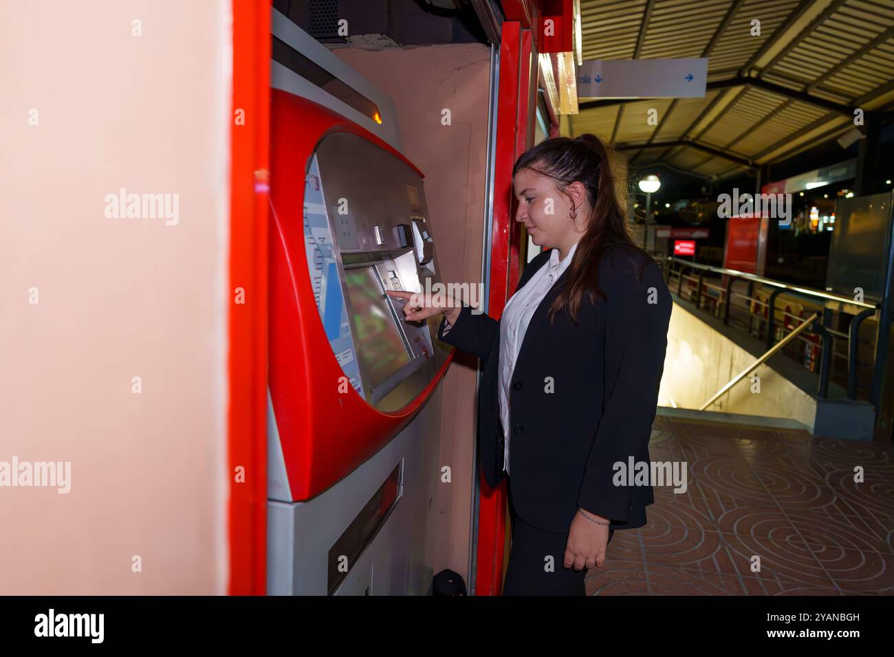 Giovane donna in abbigliamento da lavoro che utilizza la biglietteria automatica alla stazione ferroviaria durante la notte Foto Stock
