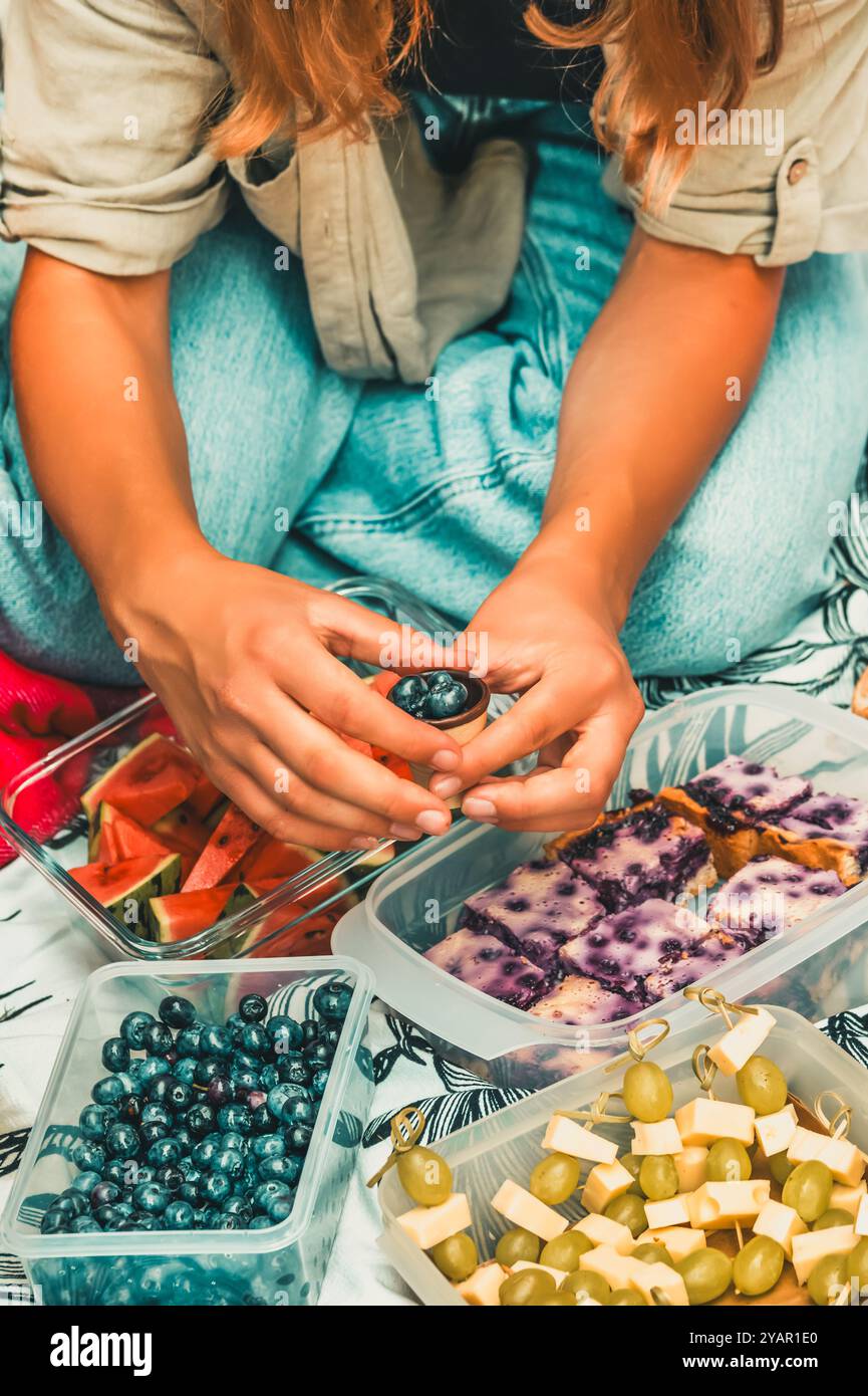 Picnic sull'erba con vari stuzzichini, mani femminili prendono mirtilli, verticale Foto Stock