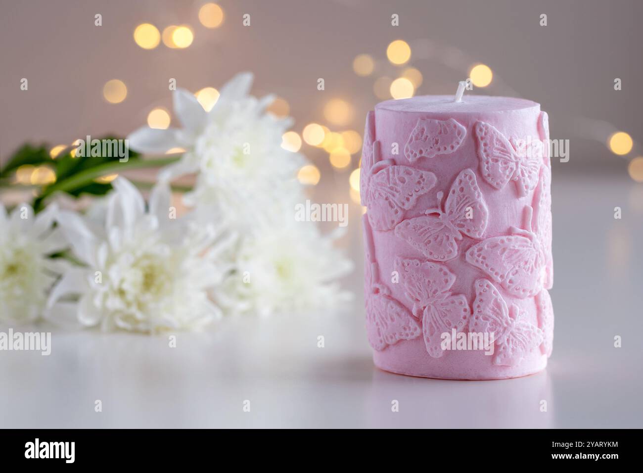 Candela decorativa rosa con motivo a farfalla. Fiori e luci sullo sfondo con spazio di copia. Arredi per la casa. Interni accoglienti. Preparazione di candele di cera di soia. Foto Stock