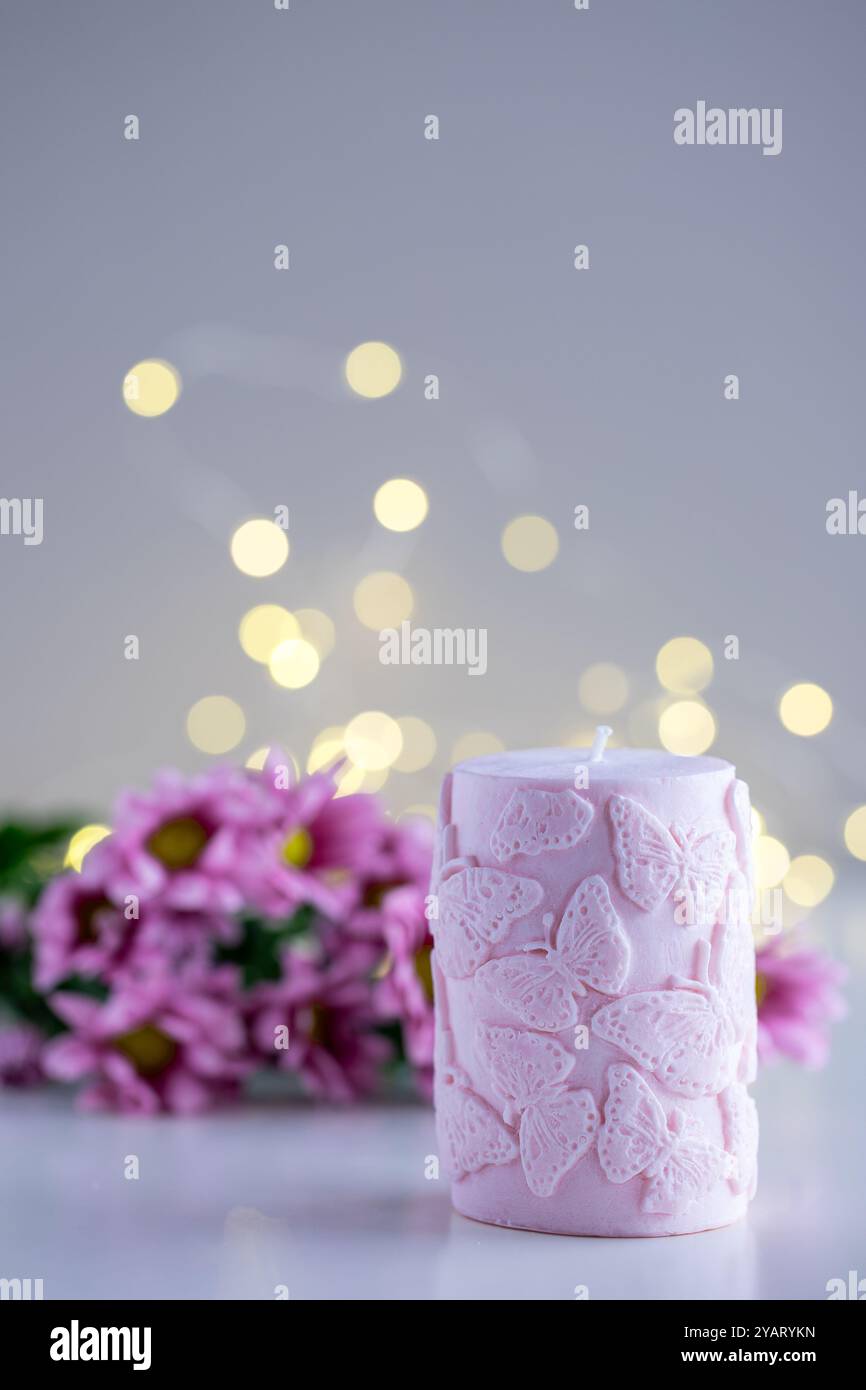 Candela decorativa rosa con motivo a farfalla. Fiori e luci su sfondo viola con spazio di copia. Arredi per la casa. Interni accoglienti. Candela di cera di soia ma Foto Stock