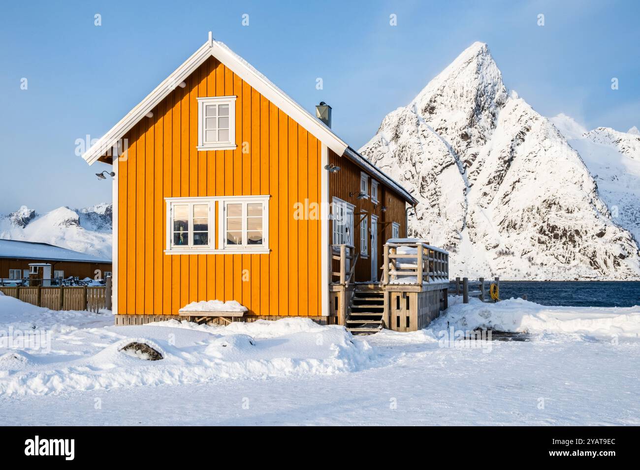 Tradizionale casa di rorbu gialla e vetta della montagna Olstind nel villaggio di pescatori di Sakrisoy sulle isole Lofoten, Norvegia. Paesaggio invernale con montagne innevate Foto Stock