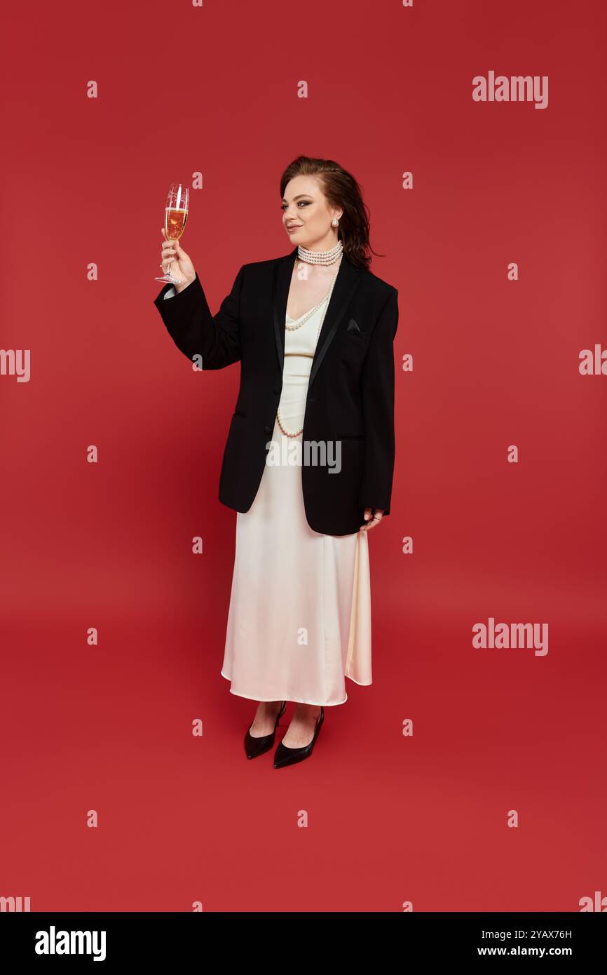Una giovane donna vestita elegantemente con un blazer nero e un lungo abito bianco solleva un bicchiere di champagne. Foto Stock
