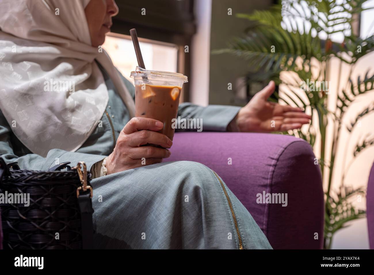 Una donna anziana che indossa un velo, si gode un rinfrescante caffè ghiacciato mentre si rilassa in un intimo caffè. L'atmosfera e' calma e invitante, raffigurante un Foto Stock