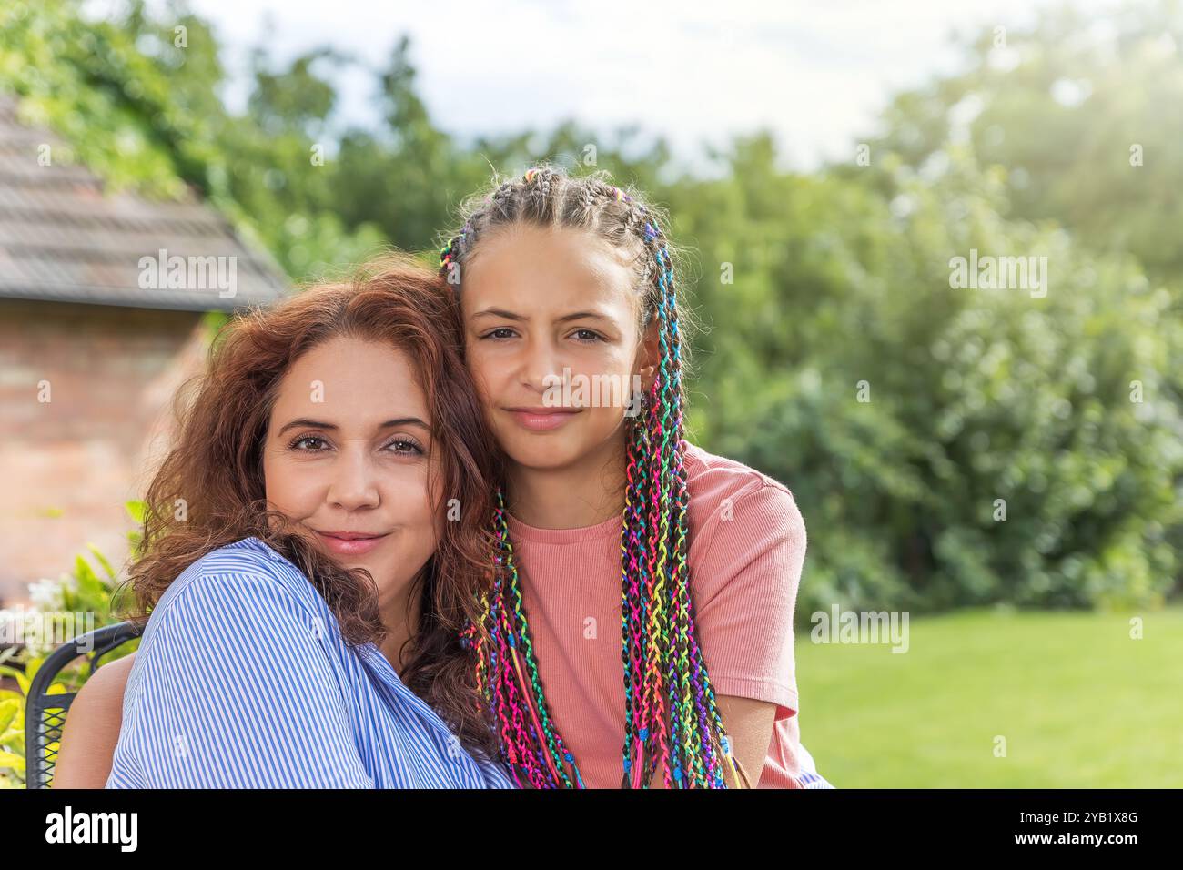 Ritratto di madre e figlia dai capelli lunghi che guardano la macchina fotografica che posa in giardino. Orizzontalmente. Foto Stock