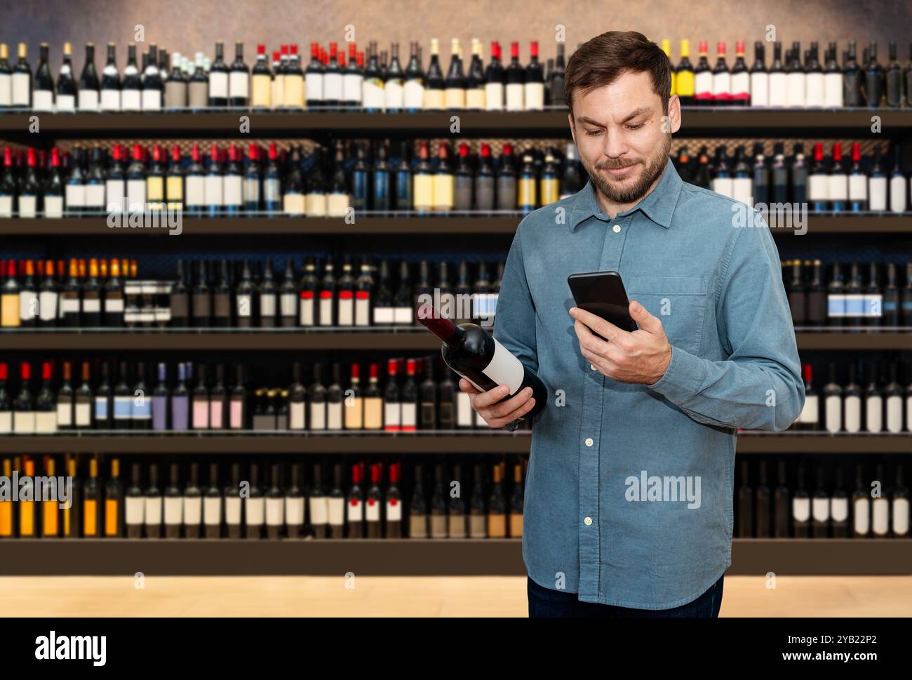 Un cliente maschio si trova in un negozio di liquori con in mano una bottiglia di vino e il suo smartphone, scansionando il codice QR per saperne di più sul prodotto. Foto Stock