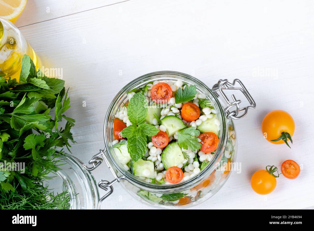 Insalata di couscous fresca in un vaso di vetro con cetrioli, pomodori, erbe aromatiche, limone con ingredienti su sfondo bianco. Insalata di tabbouleh. Vegetariano sano Foto Stock
