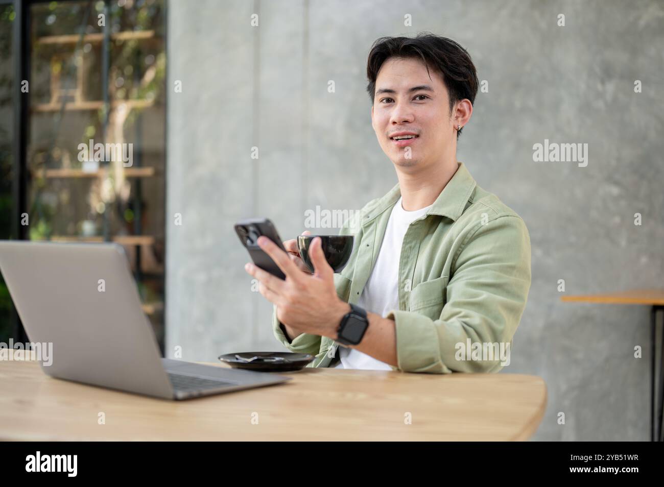 Un uomo asiatico bello e felice in abiti informali è seduto a un tavolo in una caffetteria, tenendo in mano uno smartphone e una tazza di caffè, con un computer portatile sul tavolo Foto Stock