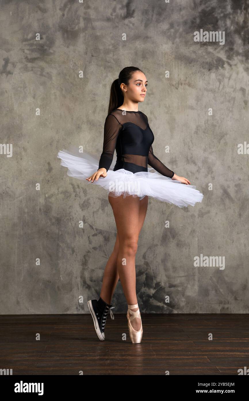 Corpo completo di giovane ballerina in tutù bianco, scarpa pointe e sneaker in piedi sui piedi mentre si pratica balletto contro il muro grungy Foto Stock